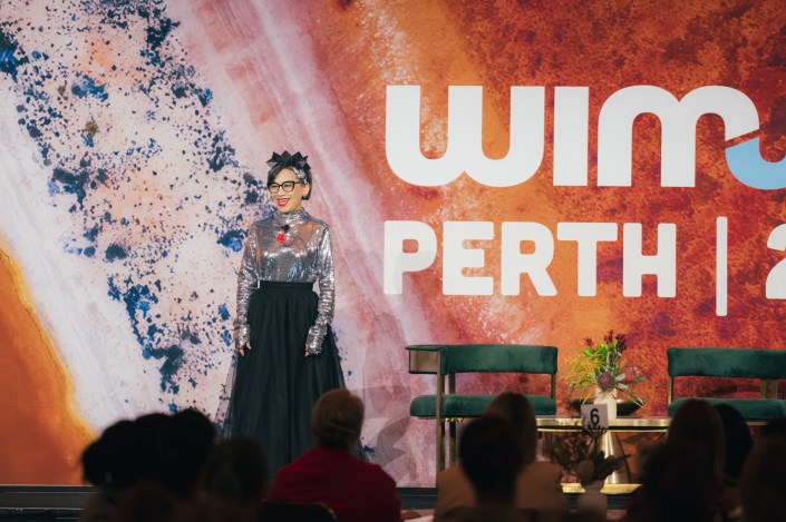 One of the speakers Rachael West coached for WIMWA 2026, Lan Tran. Woman on a stage dressed in galactic top and skirt with background reflecting the rust colours of the WA outback.